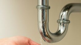 Goutte d’eau tombant d’un siphon chromé dans la main d’une femme, illustrant une fuite domestique
